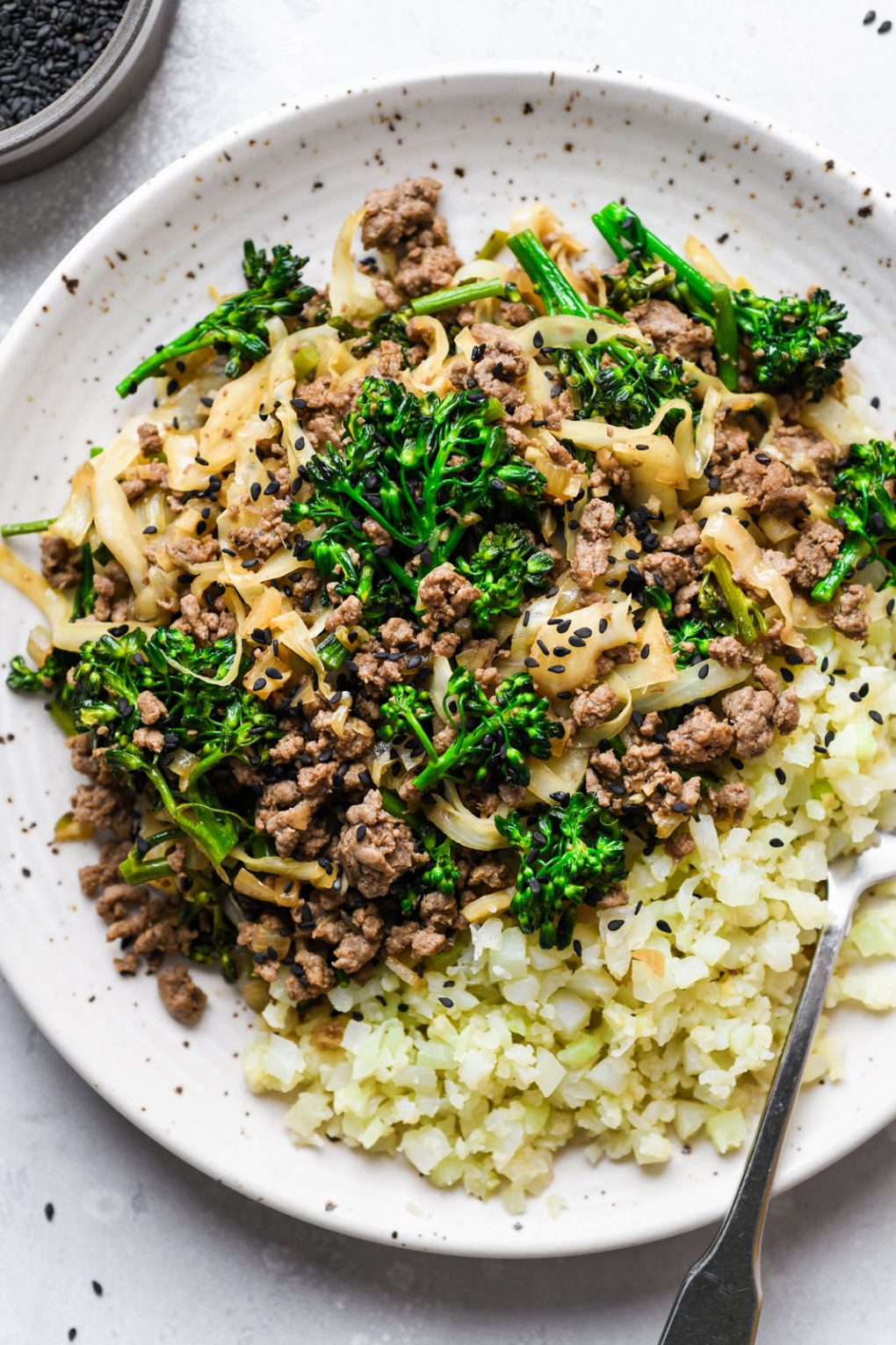 Overhead shot of a white speckled plate with ground beef and cabbage whole30 stir fry with cut up broccolini, served next to some cauliflower rice and topped with black sesame seeds. 