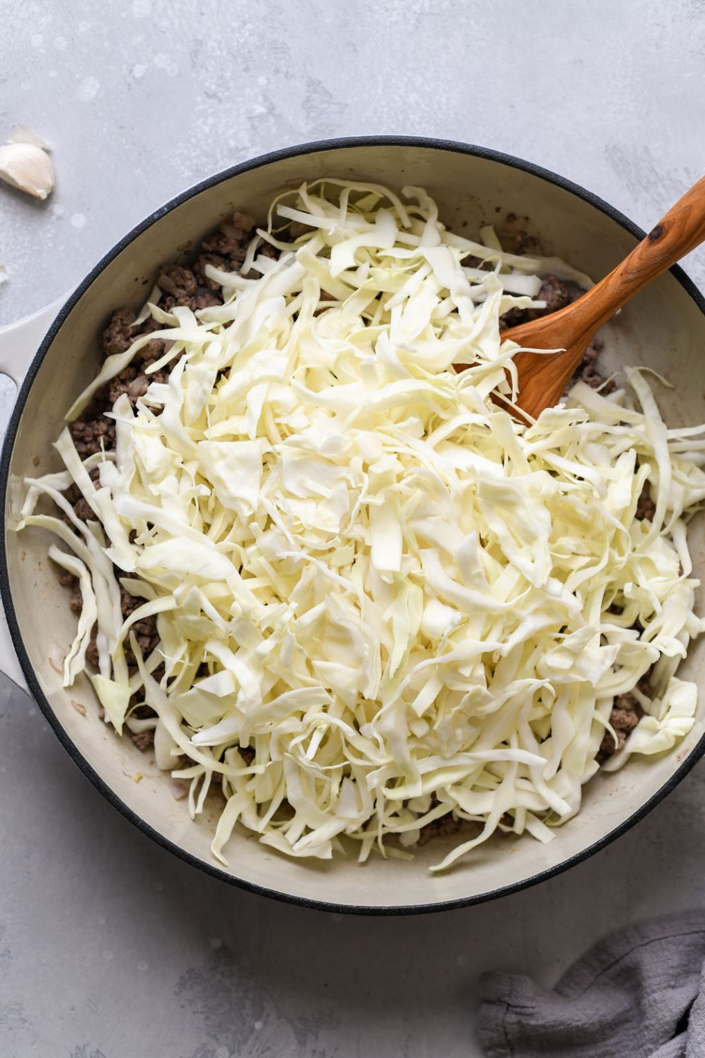 Overhead shot of a cream colored ceramic skillet filled with cooked ground beef, shallots, garlic, and piled high with fresh shredded green cabbage.