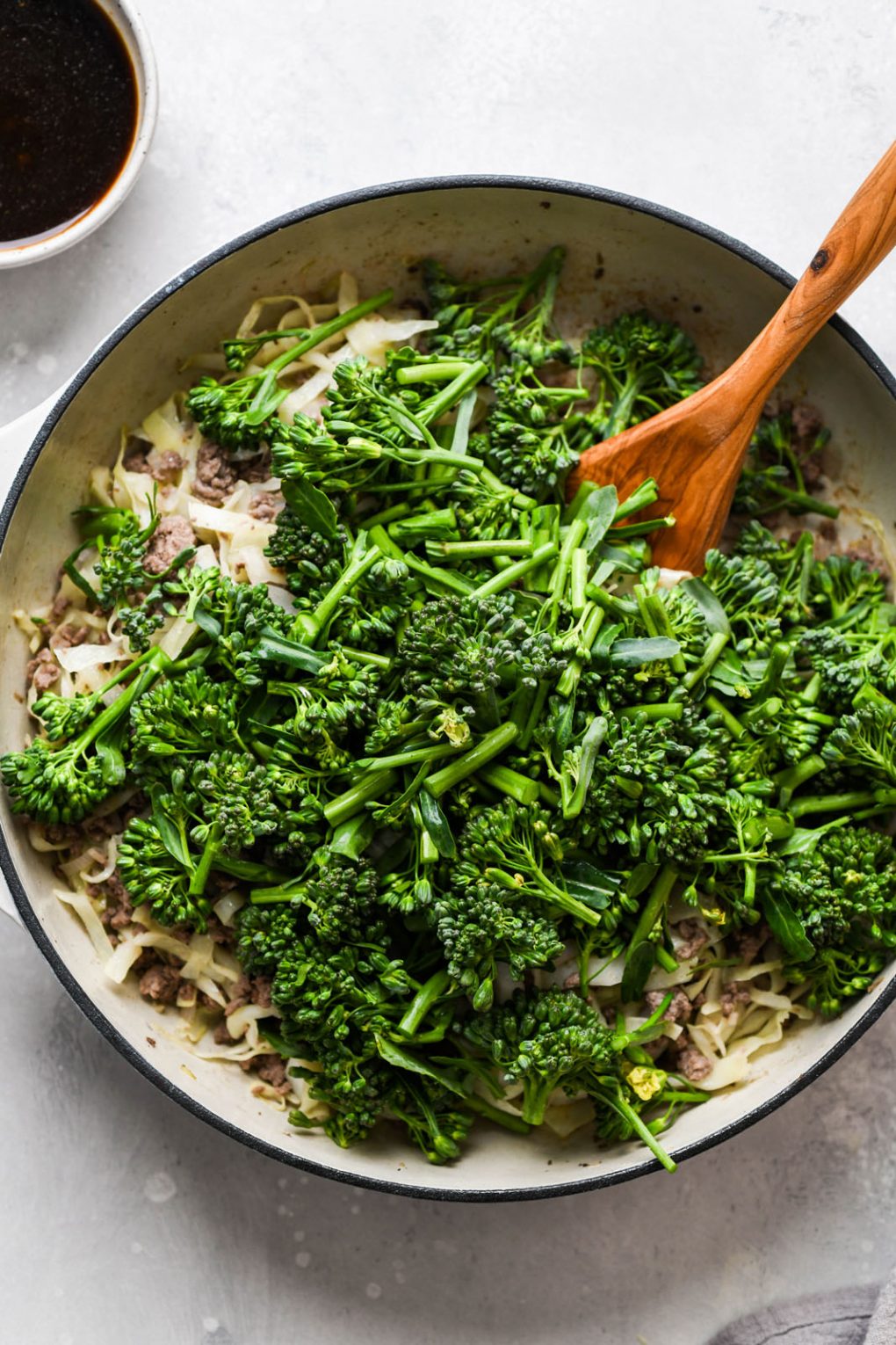 Overhead shot of a cream colored ceramic skillet filled with cooked ground beef, shallots, garlic, cooked cabbage, and piled high with fresh cut broccolini.