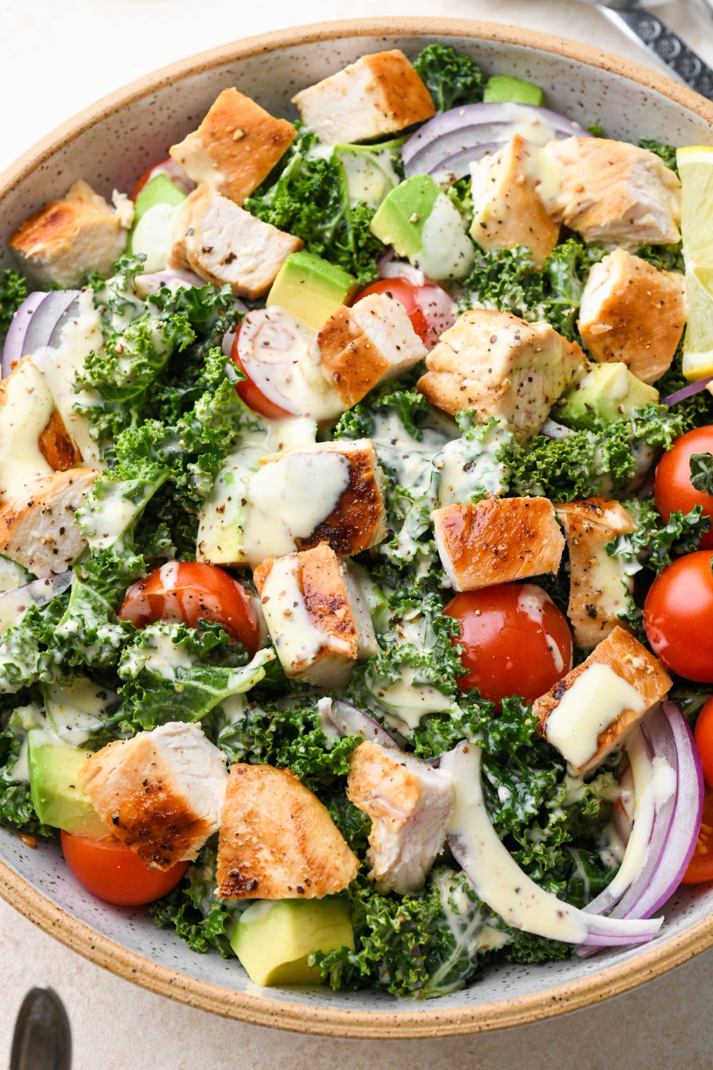 A large bowl of colorful kale chicken caesar salad with avocado, cherry tomatoes, and red onion.