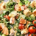 A large bowl of colorful kale chicken caesar salad with avocado, cherry tomatoes, and red onion topped with dairy free parmesan "cheese".