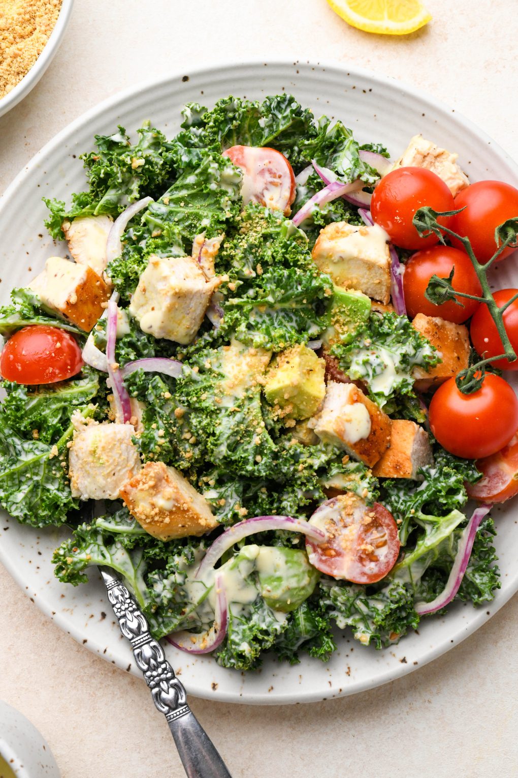 A single serving of Kale Chicken Caesar Salad on a small ceramic speckled plate, on a light colored cream background.
