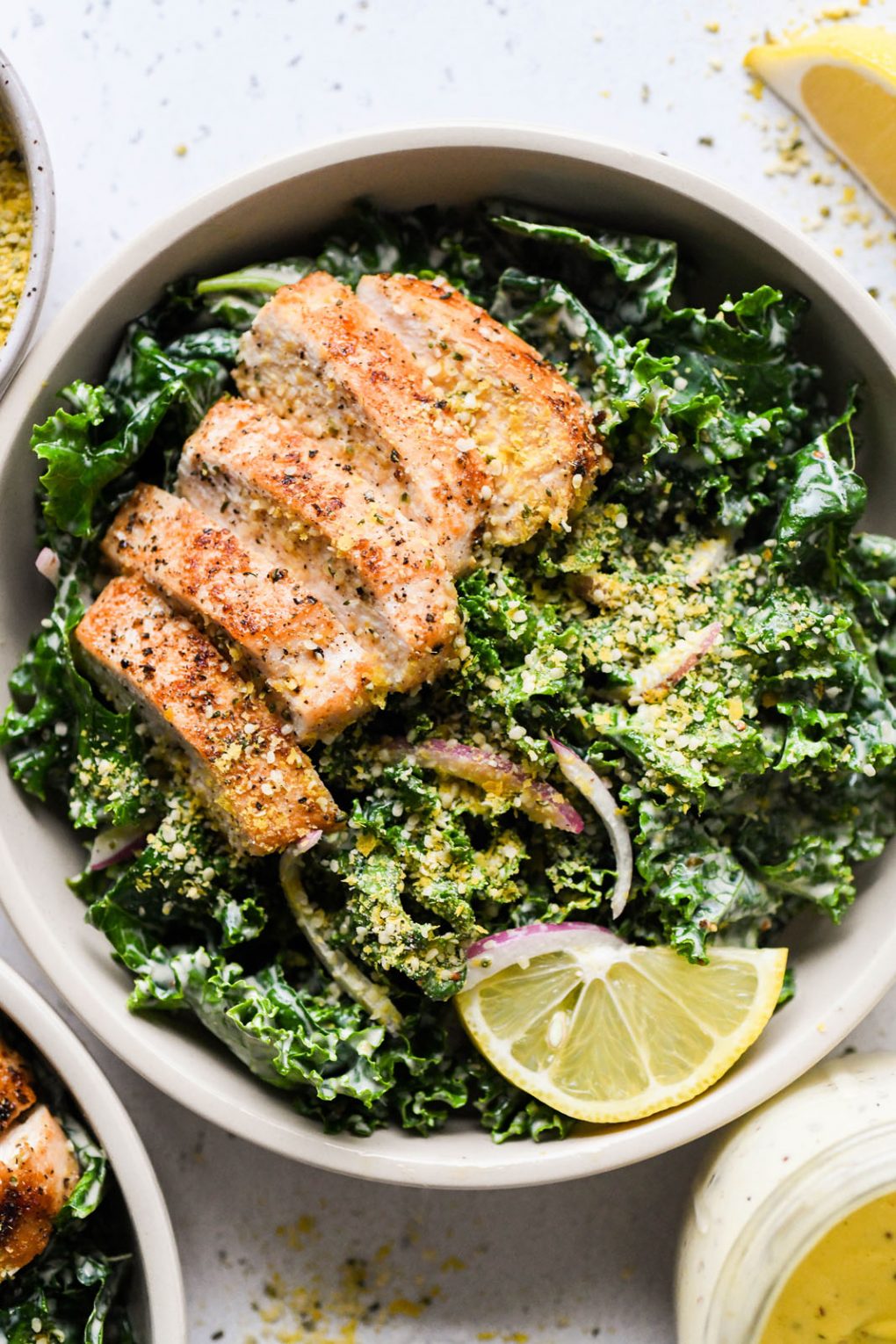 Overhead shot of a large white bowl filled with a hearty kale chicken caesar salad. Topped with a dairy free hemp heart parmesan cheese. On a light background next to a lemon wedge and a small glimpse of the jar of caesar salad dressing.