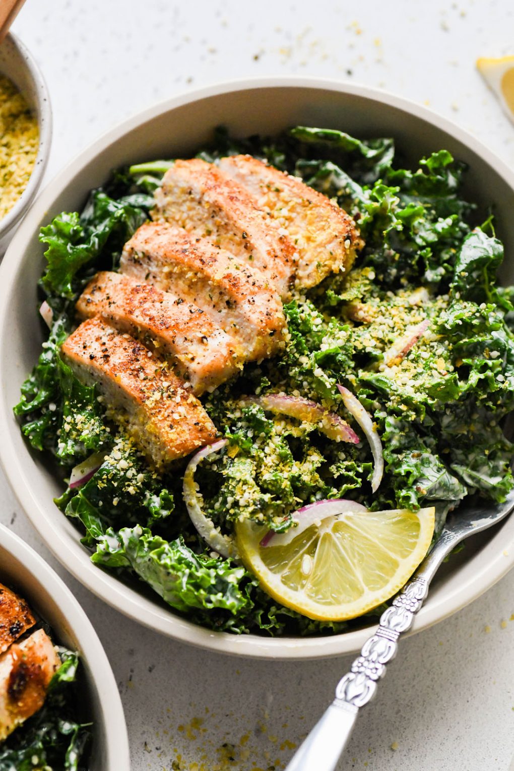 Overhead shot of a large white bowl filled with a hearty kale chicken caesar salad. Topped with a dairy free hemp heart parmesan cheese. On a light background next to a lemon wedge and a small glimpse of the jar of caesar salad dressing.