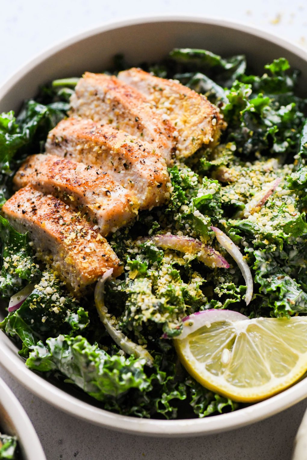 Super close up shot of a large white bowl filled with a hearty kale chicken caesar salad. Topped with a dairy free hemp heart parmesan cheese.
