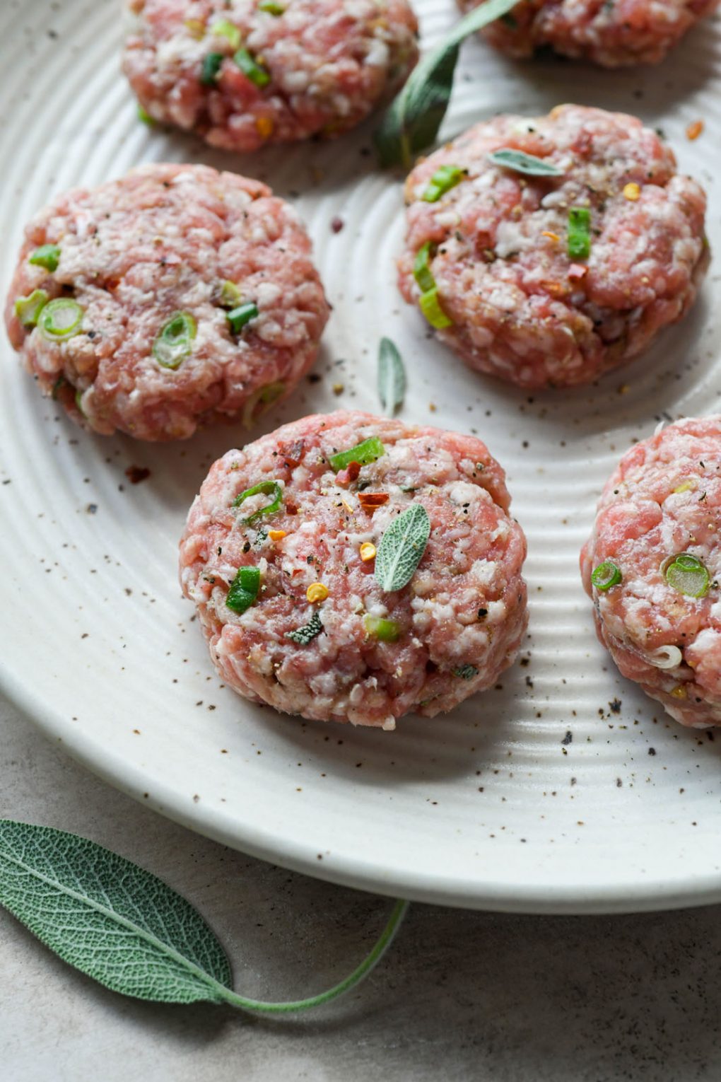 Close up 45 degree angle shot of several raw Whole30 pork sausage patties scattered with fresh herbs. 