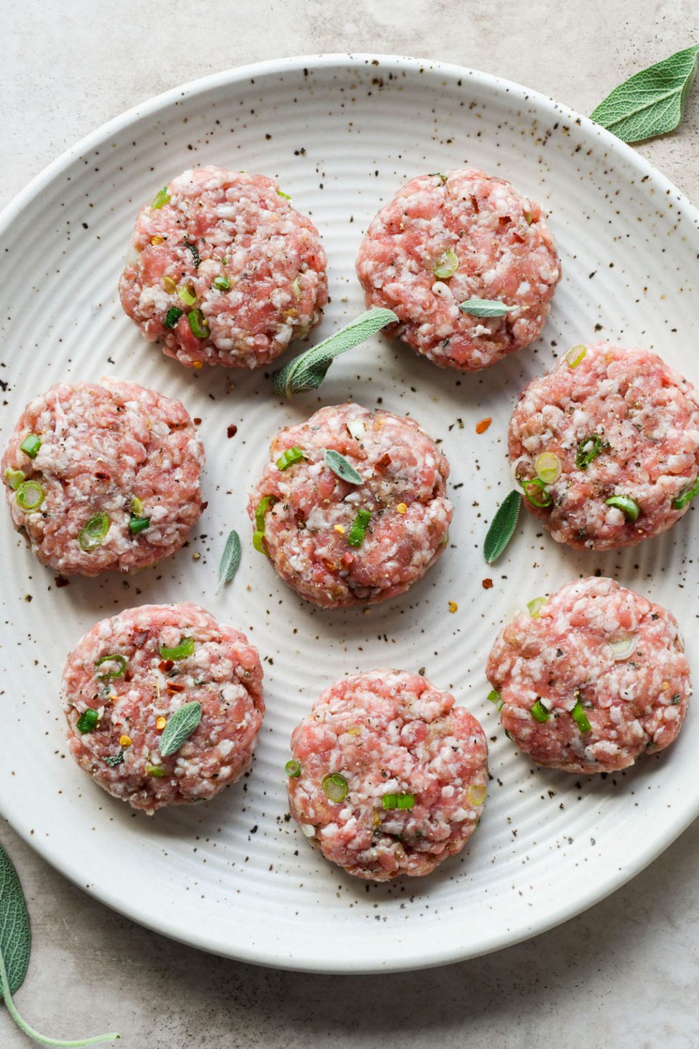 Overhead shot of several raw Whole30 pork sausage patties scattered with fresh herbs. 