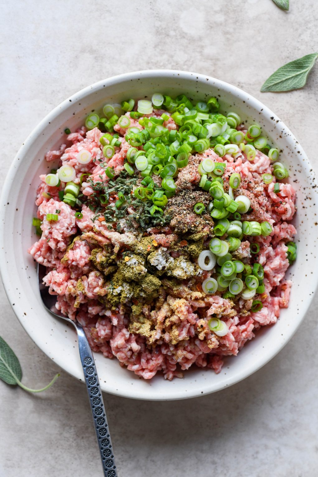 Overhead shot of a light colored speckled bowl with whole30 pork sausage ingredients - ground pork, spices, salt, pepper, sliced green onions, chili flakes, and chopped fresh sage. On a creamy colored background with an antique spoon tucked into the bowl. 