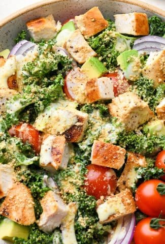 A large bowl of colorful kale chicken caesar salad with avocado, cherry tomatoes, and red onion topped with dairy free parmesan "cheese".