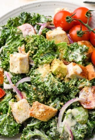 A single serving of Kale Chicken Caesar Salad on a small ceramic speckled plate, on a light colored cream background.