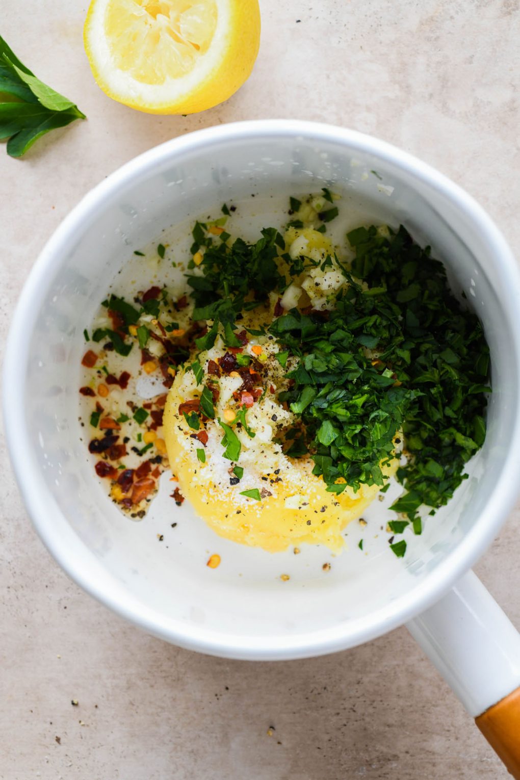 A small white sauce pan with the ingredients for garlic herb ghee - ghee, chili flakes, chopped parsley, garlic, salt and pepper. 