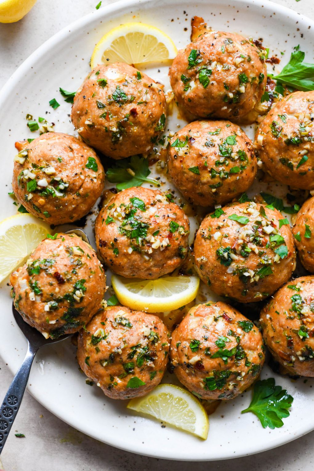 A large plate filled with large glistening herb flecked turkey meatballs drizzled with golden colored ghee and next to a few lemon slices and parsley leaves. One meatball is cut in half to reveal the interior. On a light colored speckled plate with a spoon slanted off the plate holding one meatball. 