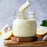 A spoon holds creamy Caesar dressing above a glass jar filled with the same dressing. Lemon wedges, garlic cloves, and parsley are blurred in the background, all set on a wooden surface.