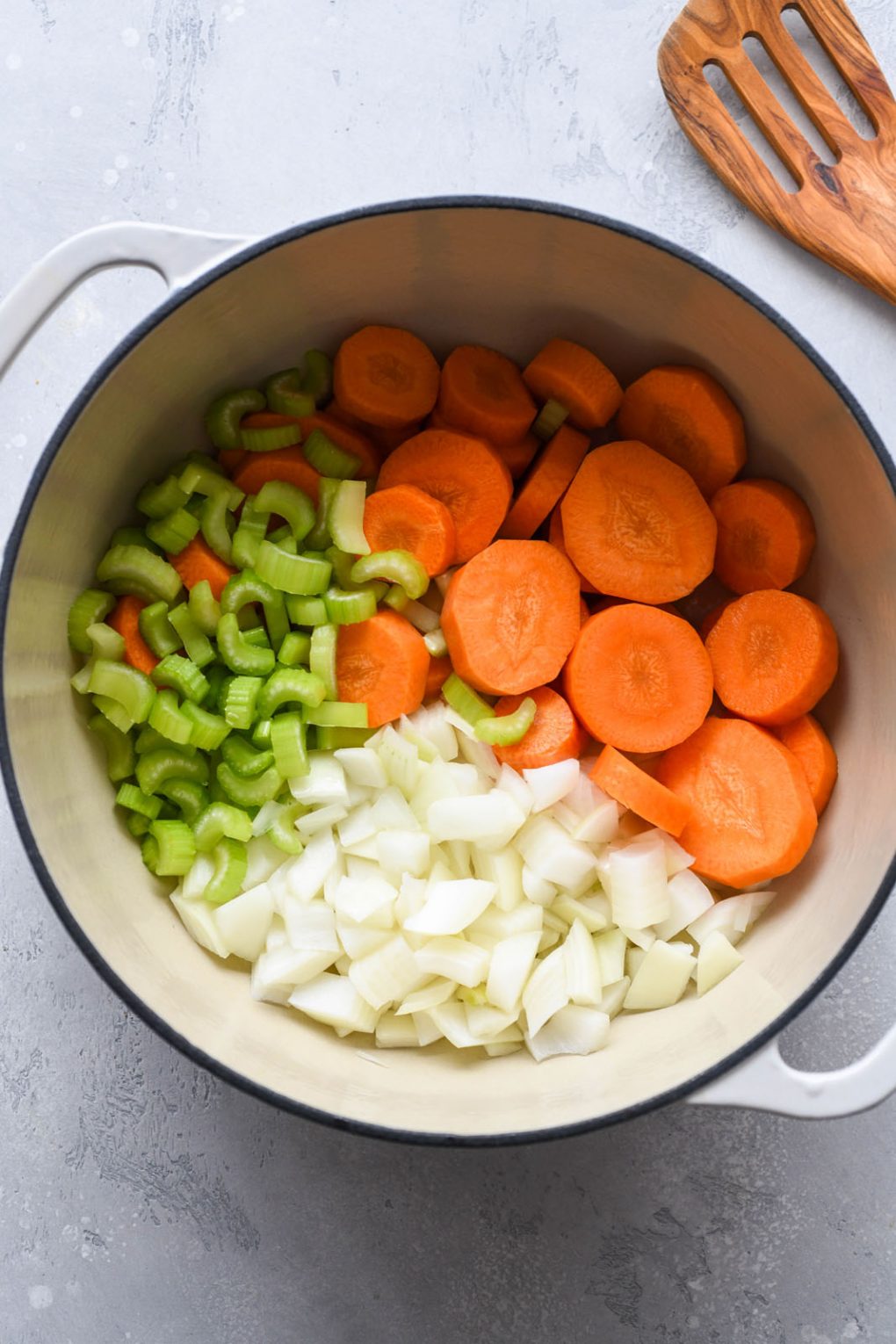 Overhead image of onion, celery, and sliced carrots, in a large white ceramic soup pot. 