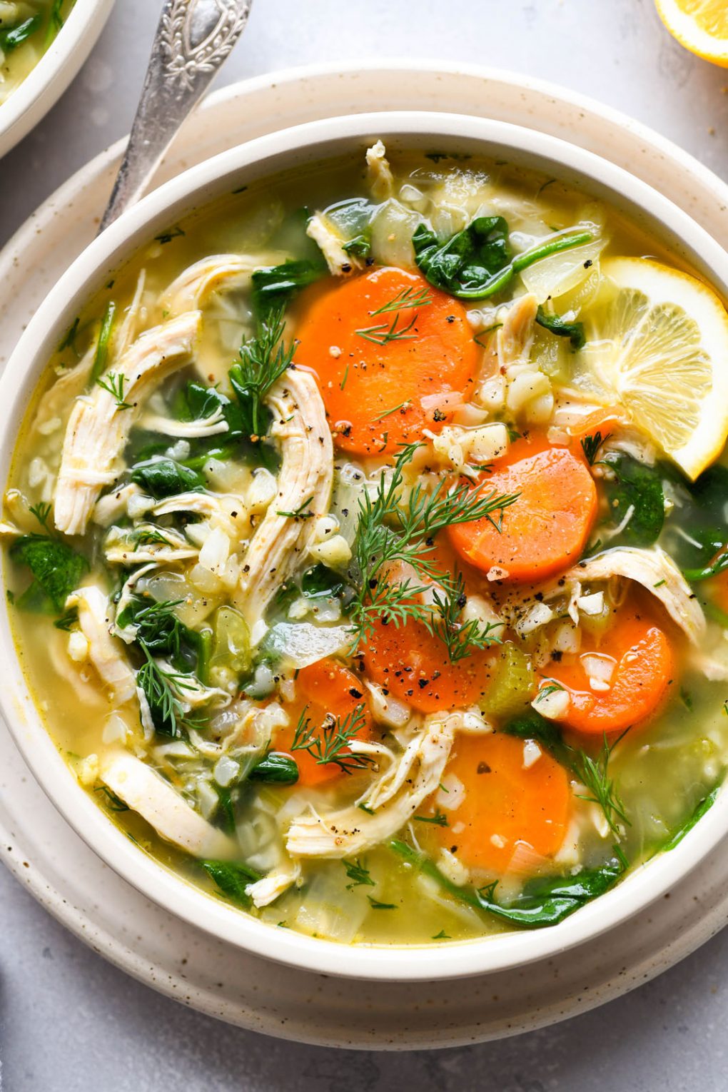 Overhead bright and glowing image of nourishing chicken and cauliflower rice soup in a shallow white bowl, on a light background surrounded by cut lemon and scattered fresh herbs. 
