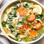 A bowl of chicken and cauliflower rice soup with shredded chicken, sliced carrots, spinach, fresh dill, and a lemon wedge, served with a spoon on a plate.