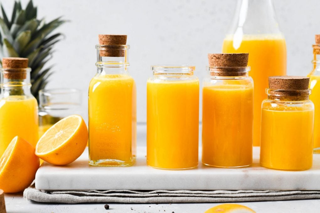Straight on photograph of various sized small jars filled with bright yellow immune boosting wellness shots. On a light background next to the top of a pineapple, cut lemon, and some scattered black peppercorns.