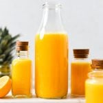 A glass bottle filled with citrus juice surrounded by pineapple, sliced oranges, and ginger.
