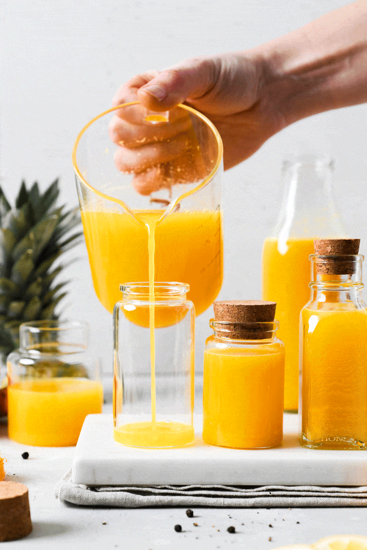 Straight on moving GIF of a measuring glass pouring juice into a small glass container, next to various sized small jars filled with bright yellow immune boosting wellness shots. On a light background next to the top of a pineapple, cut lemon, and some scattered black peppercorns.