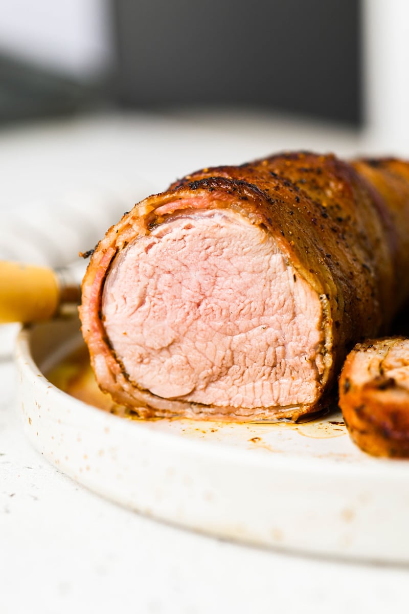 Close up image of a bacon wrapped pork tenderloin cut into so you can see the juicy, perfectly cooked meat. 