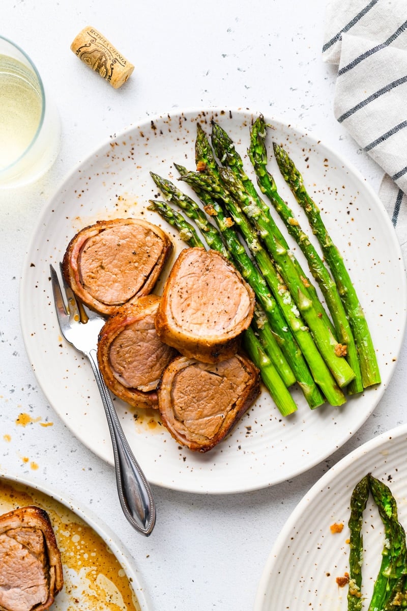 Overhead image of sliced bacon wrapped pork tenderloin on a white speckled plate with roasted asparagus. On a light colored background, next to a striped towel, a glass of white wine, and a wine cork.