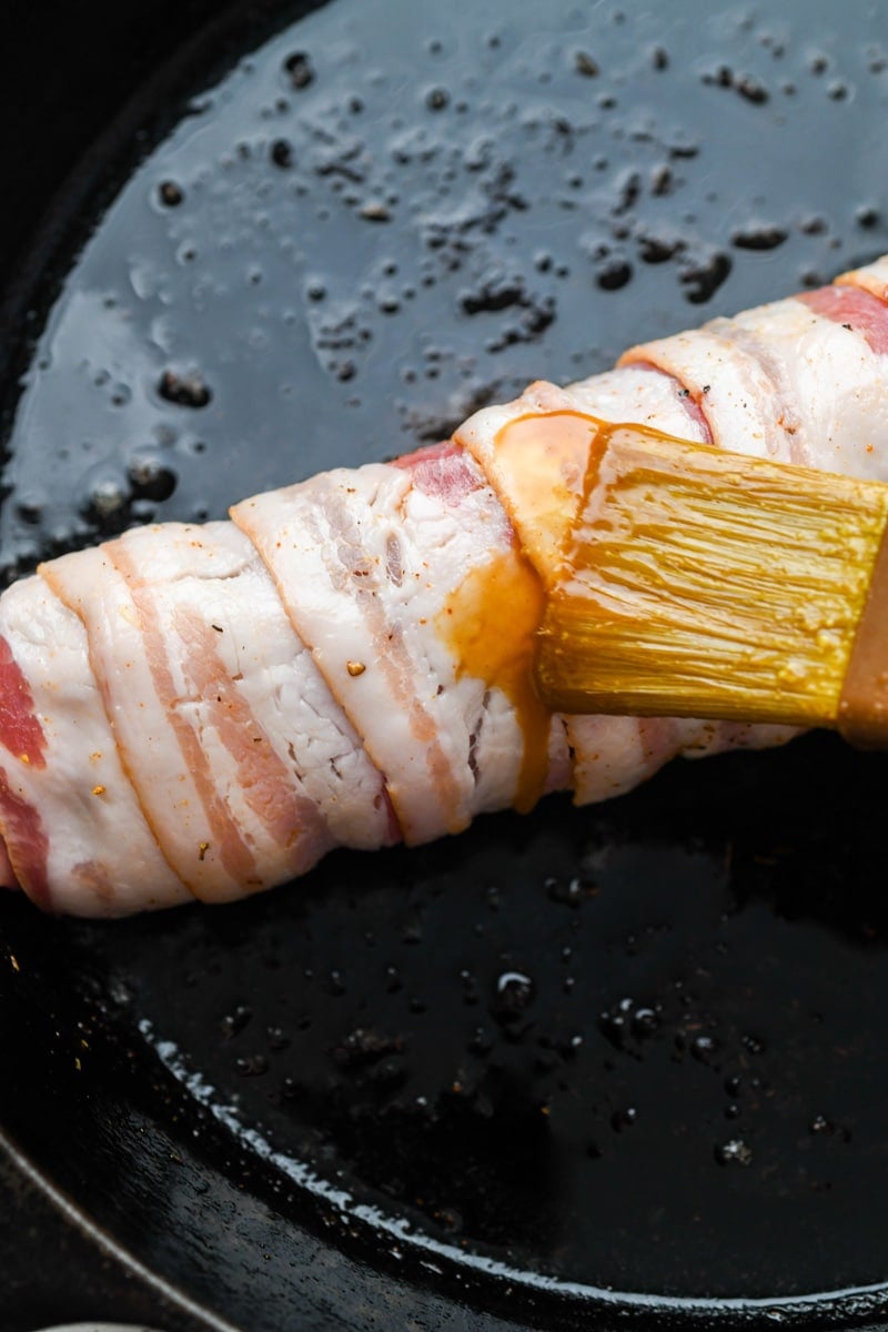 Close up shot of a bacon wrapped pork tenderloin in a cast iron skillet. A pastry brush brushing on the coconut amino mixture.