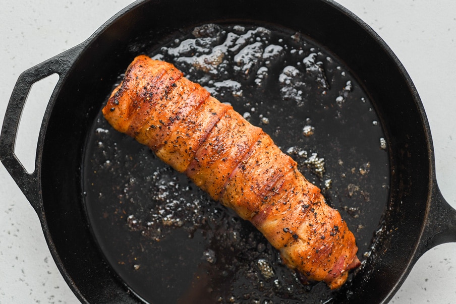 Overhead image of a roasted bacon wrapped pork tenderloin brushed with a coconut amino glaze, in a cast iron skillet.