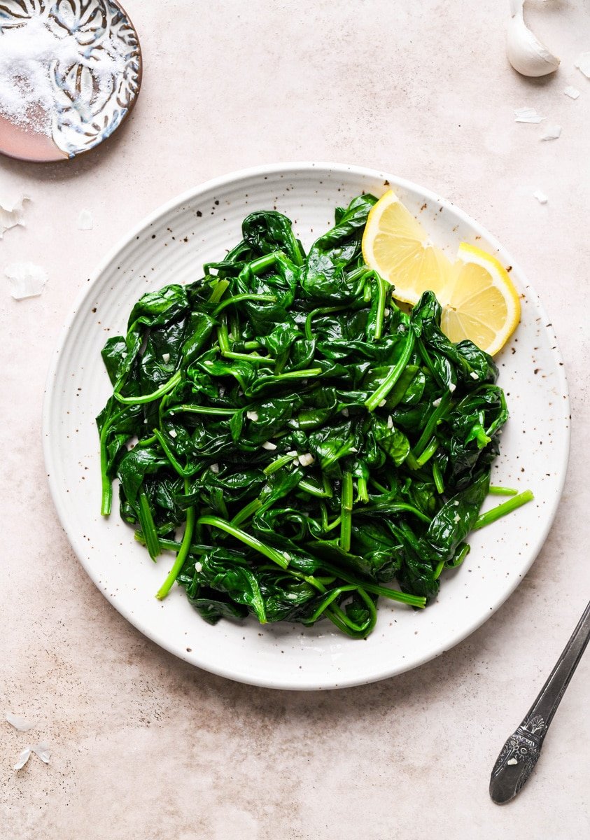 Overhead shot of a large white speckled plate of garlic sauteed spinach. Next to a few lemon wedges. On a speckled tan background, next to a small dish of salt.