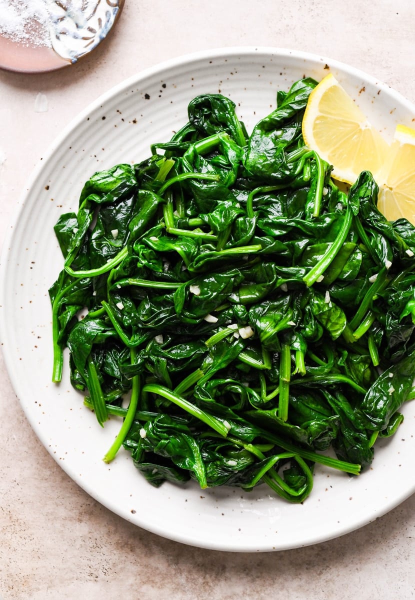 Close up shot of a large white speckled plate of garlic sauteed spinach. Next to a few lemon wedges. On a speckled tan background, next to a small dish of salt.