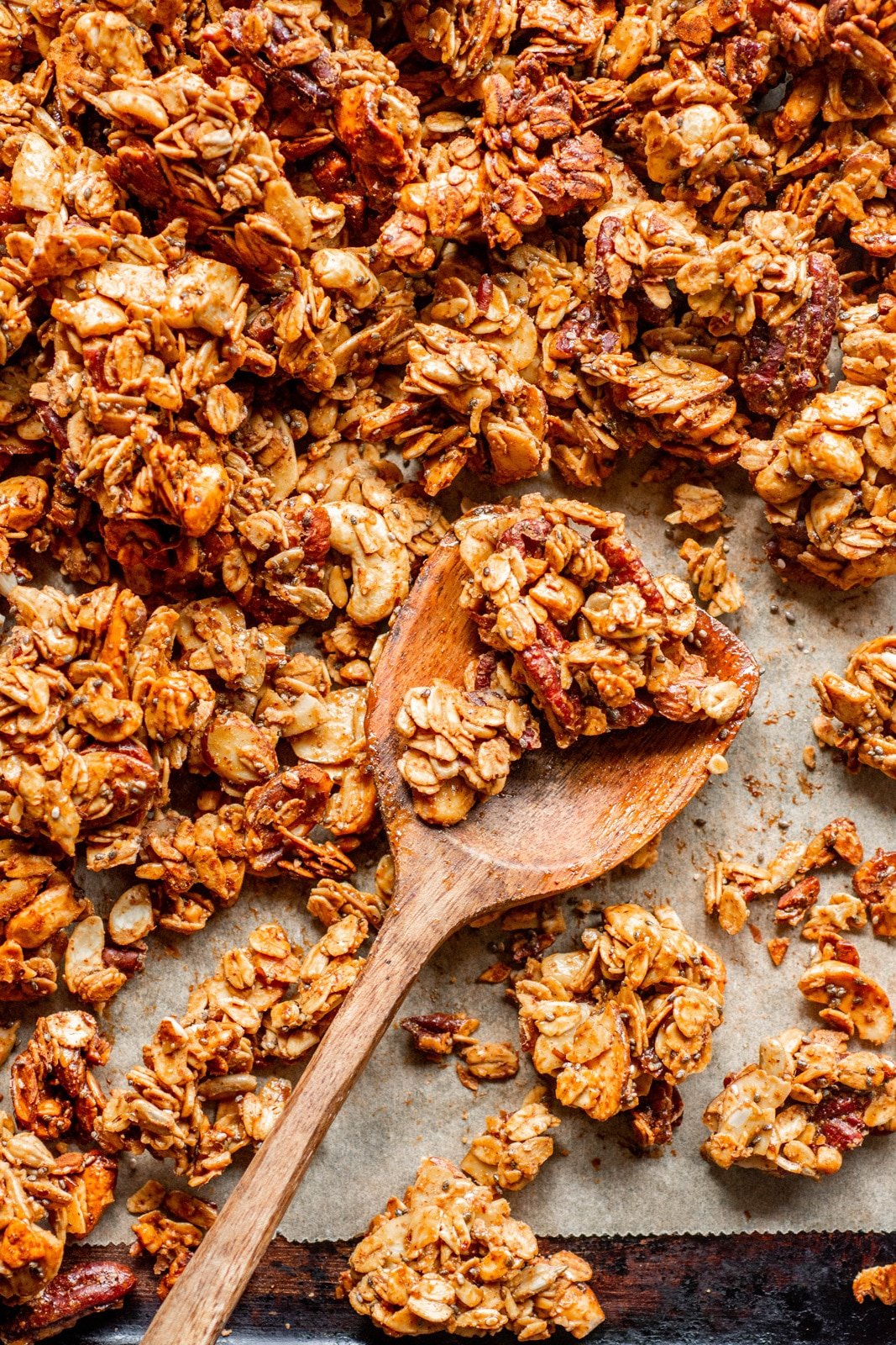 Overhead shot of a wooden spoon holding a few crunchy granola clusters, on a parchment lined baking sheet.