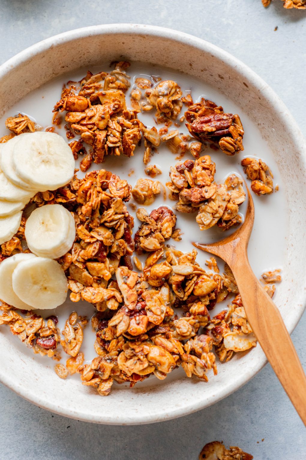 Overhead image of a white bowl on a light background, filled with chunky gluten free granola, almond milk, and sliced banana. With a wooden spoon inside the bowl. 