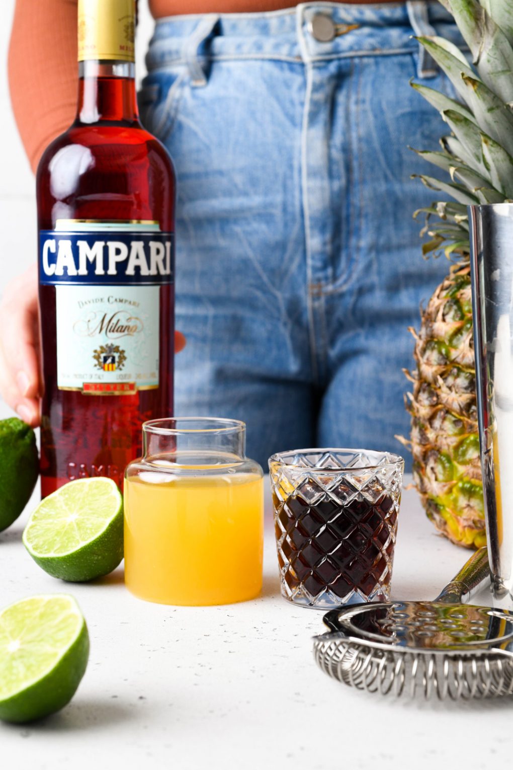 Straight on shot of the ingredients for a tropical jungle bird cocktail. A bottle of campari, a small jar of pineapple juice, a shot glass with dark rum, and some cut limes. On a white background.