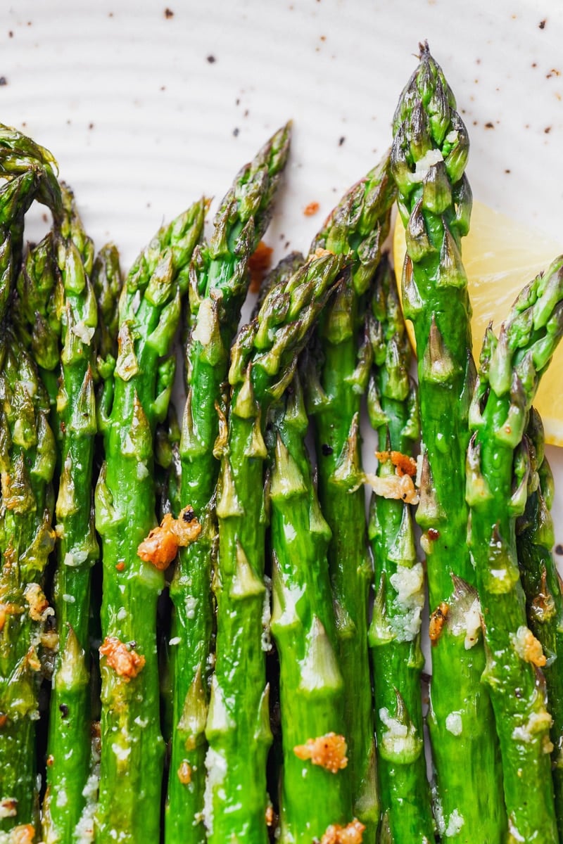 Super close up shot of tender and caramelized bright green lemon garlic asparagus spears. On a large white speckled plate, next to a slice of lemon. 