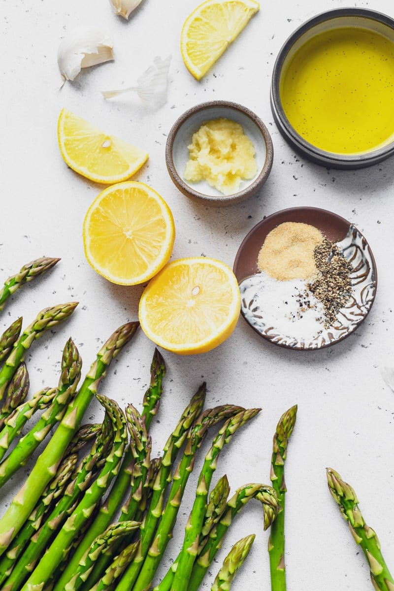 An overhead shot of scattered ingredients needed to make simple lemon garlic roasted asparagus. Asparagus spears, cut lemon, a small dish with grated garlic, another small dish with garlic powder, salt and pepper, and a bowl of olive oil. On a light colored speckled background. 