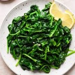 A white plate with sautéed spinach garnished with two lemon wedges, placed on a light-colored surface.