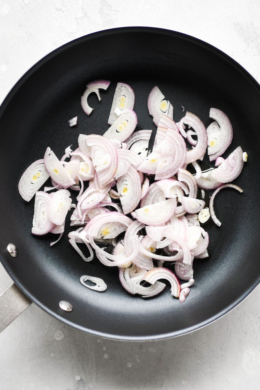 Overhead shot of a medium sized non stick skillet filled with raw thinly sliced shallots. On a light colored background. 
