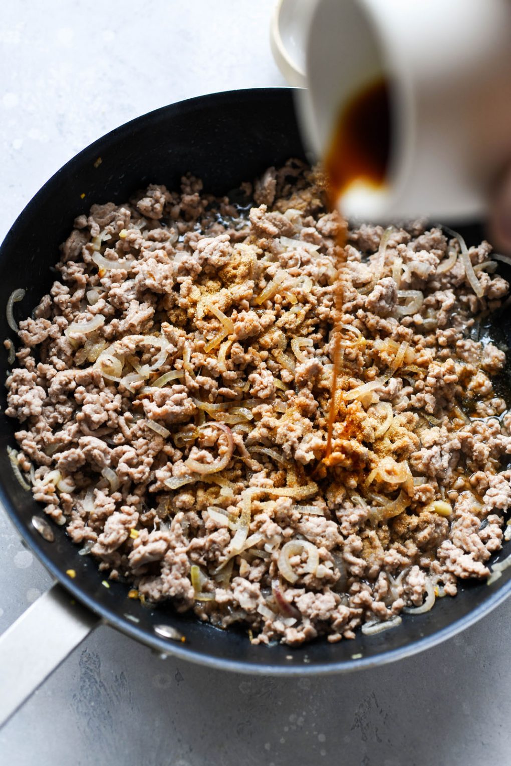Close up shot of cooked ground pork, with a hand pouring coconut aminos into the pan. 