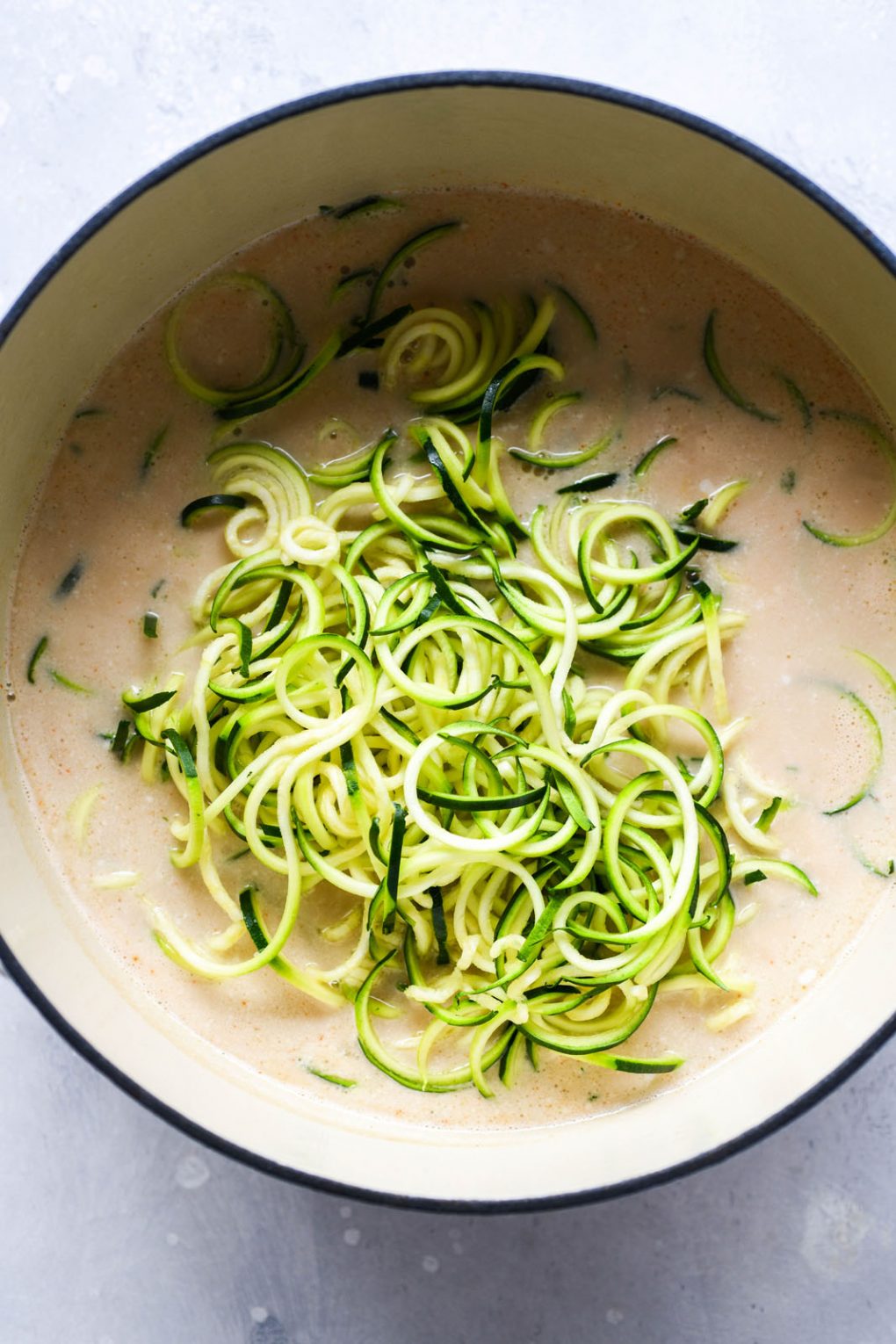 Overhead shot of a large white ceramic soup pot filled with coconut broth, and bright green raw zucchini noodles.