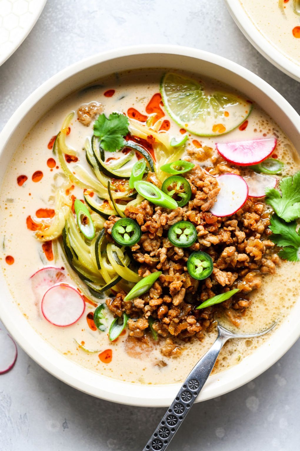 Overhead shot of a bowl of zucchini noodle soup with crispy pork and a coconut broth. Topped with thinly sliced radishes, green onions, jalapeno, cilantro, chili oil, and lime wedges. In a light colored bowl on a white background, with a spoon dipped into the side of the bowl. 