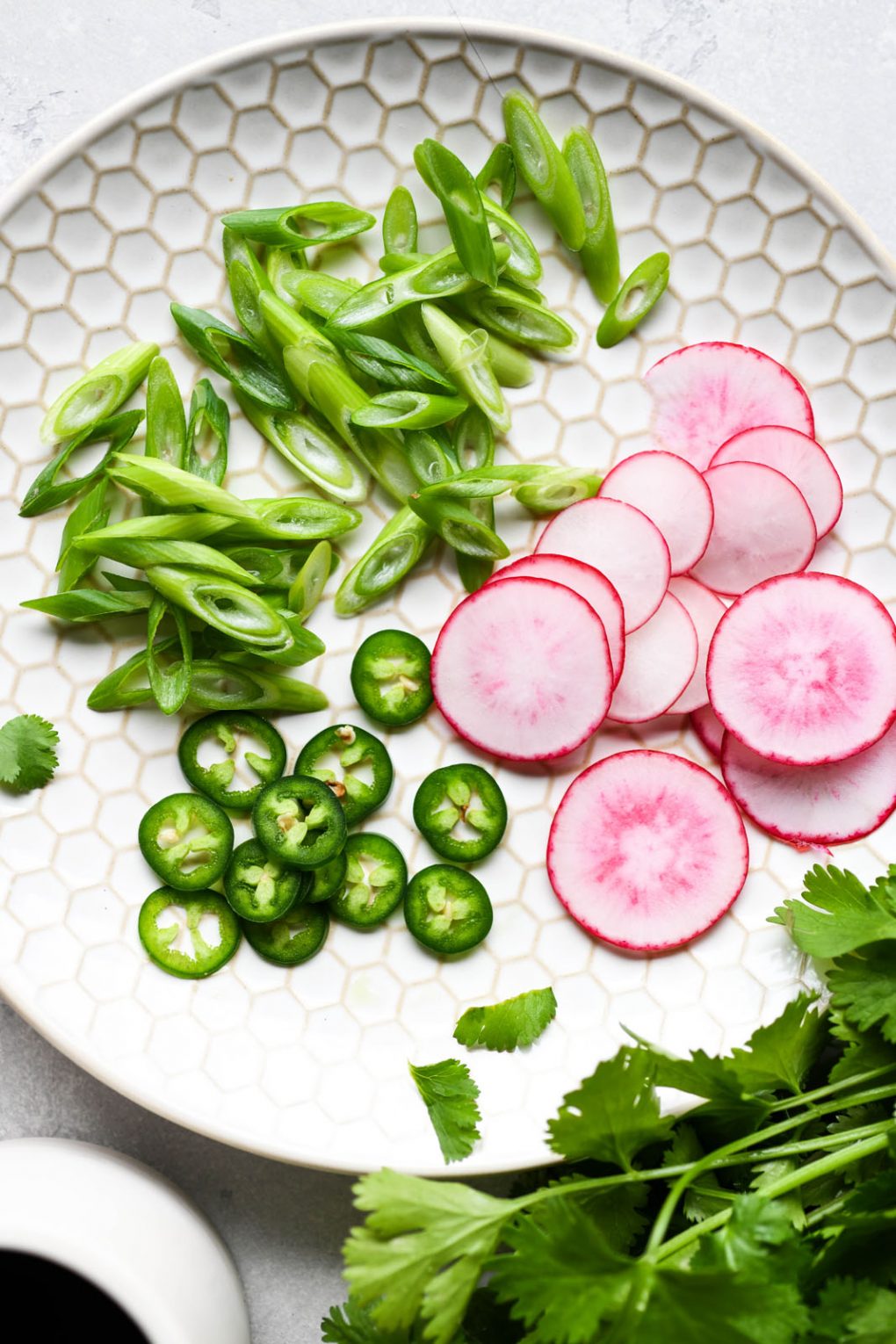 Super close up image of soup garnishes on a light colored textured plate. Thinly sliced radishes, jalapeno, green onions, and cilantro peeking into the corner of the frame. 