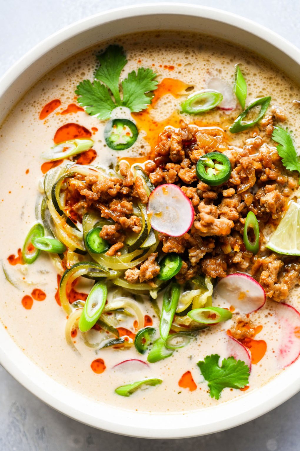 Overhead shot of a bowl of zucchini noodle soup with crispy pork and a coconut broth. Topped with thinly sliced radishes, green onions, jalapeno, cilantro, and chili oil. In a light colored bowl on a white background.