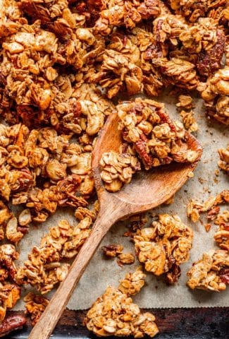 Overhead shot of a wooden spoon holding a few crunchy granola clusters, on a parchment lined baking sheet.