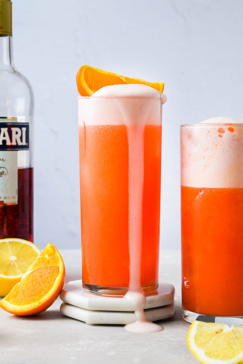 Straight on image of two side by side orange campari vodka sour cocktails, the centered cocktail with an orange wedge on the top of the drink and egg white foam flowing over the side of the high ball glass. On a light background next to some scattered orange and lemon wedges, with a bottle of Campari in the background.