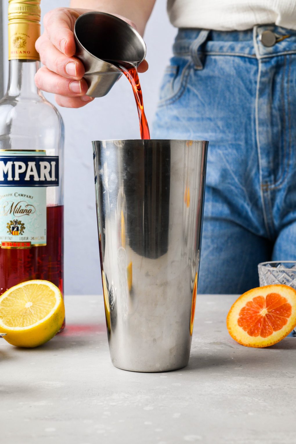 A woman standing behind a light colored counter pouring campari into a large silver cocktail shaker.