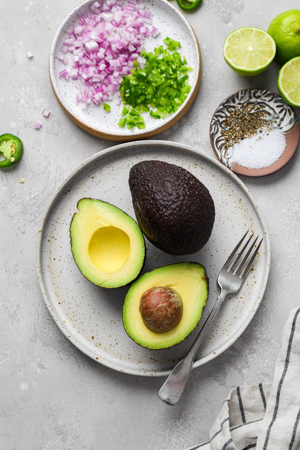 Image of ingredients used to make easy guacamole recipe. A plate with 2 avocados - one cut in half, a smaller plate with finely diced red onion, jalapeno, some cut limes, and a smaller plate with salt and pepper. 
