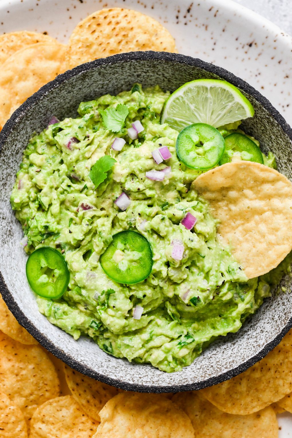 Overhead shot of a small bowl of bright green guacamole with a chip tucked into the side of the bowl. Guacamole is topped with some finely diced onion, thinly sliced jalapeño, and cilantro leaves, surrounded by round yellow tortilla chips. 