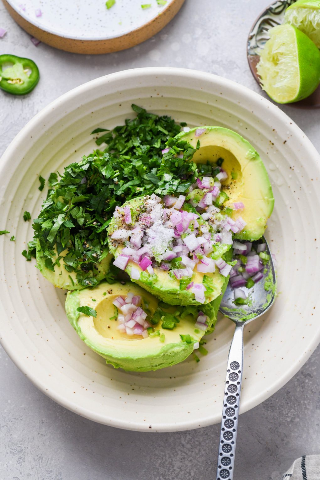 Image of a large cream colored speckled bowl with ingredients for easy guacamole - avocado scooped out of the skin, diced red onion, jalapeno, cilantro, and salt and pepper. On a light grey background. 
