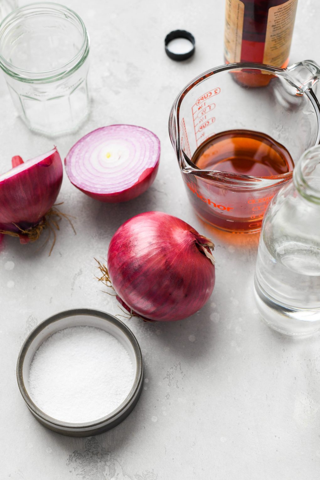 Overhead shot of ingredients for quick pickled red onions - a large red onion next to another large red onion cut in half, a measuring cup with red wine vinegar, a small dish of kosher salt, and a jar of water.