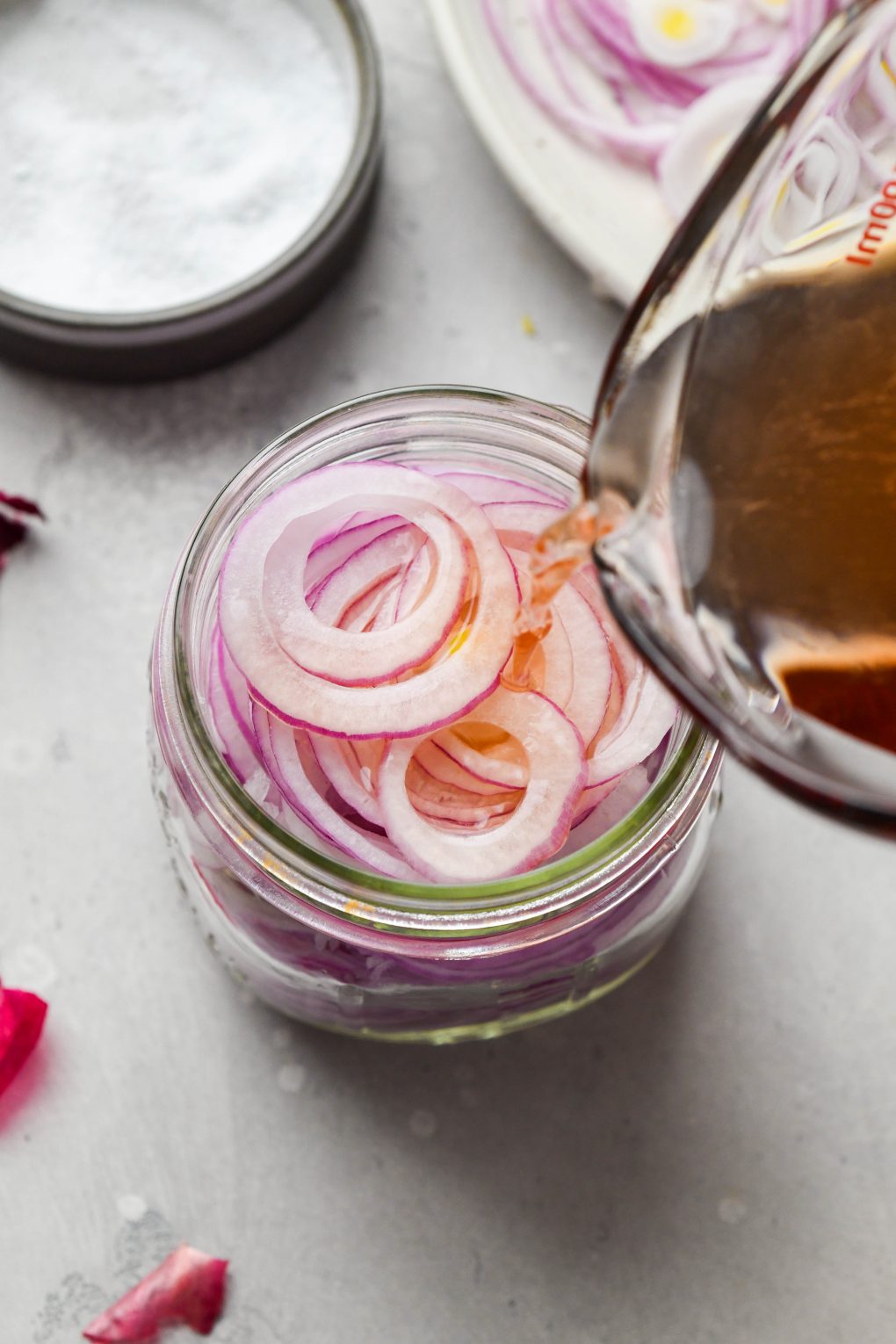 A 45 degree angle shot of a medium sized glass jar filled with bright pink thinly sliced red onion and a glass measuring cup pouring pickling liquid into the jar.