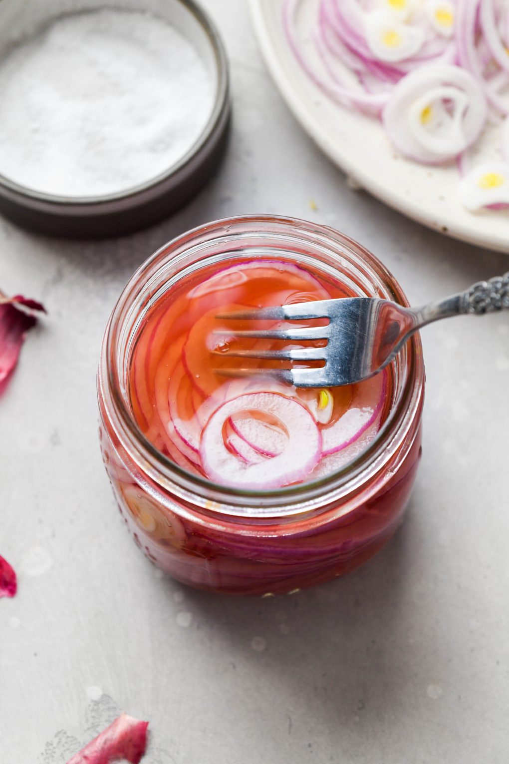 A 45 degree angle shot of a medium sized glass jar filled with bright pink thinly sliced red onion. On a light grey background with a fork pressing the red onion slices into the liquid and scattered red onion skins.